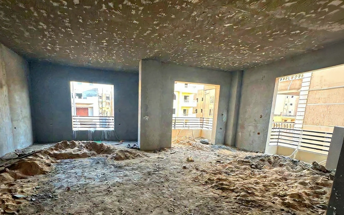 Та же комната до ремонта: голые бетонные стены и потолок, песок на черновом полу, пустые проёмы окон и балкона